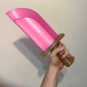 May include: A pink toy sword with a brown handle and crossguard. The blade is a vibrant pink, and the handle is a contrasting brown. The sword is held in a hand against a plain background.
