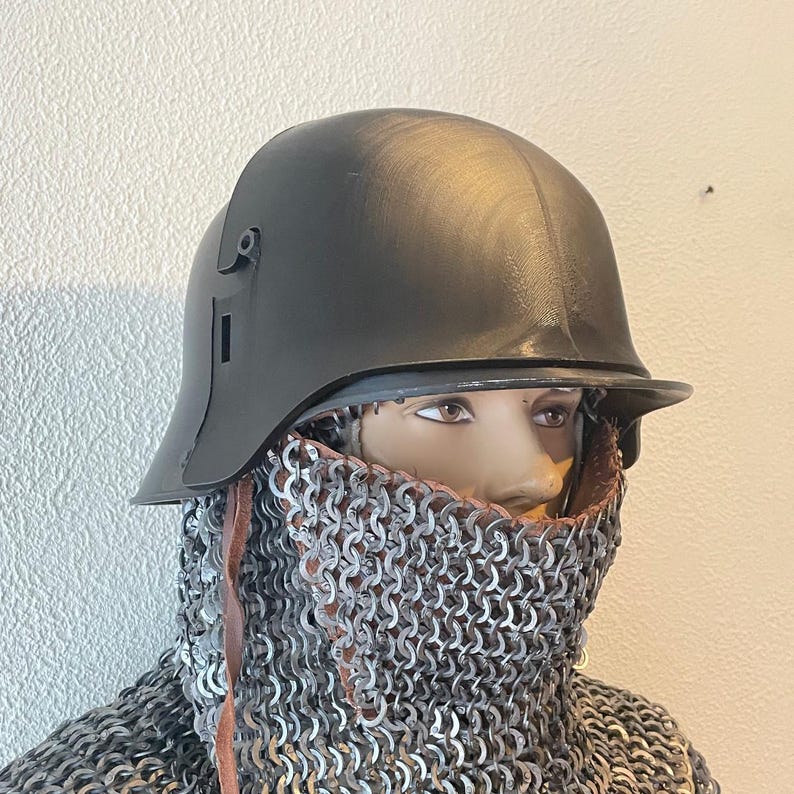 May include: A dark gray helmet with a visor and side details, worn over a chainmail coif. The helmet is displayed on a mannequin head. The chainmail covers the neck and shoulders, with leather straps visible.