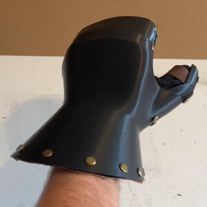 May include: A black gauntlet glove featuring a smooth, curved outer shell with a flared cuff. The piece is constructed with metallic brass-coloured rivets along the edges and includes a brown leather strap detail visible near the thumb area.