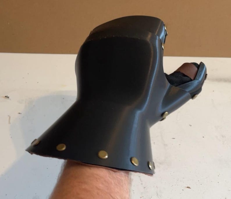 May include: A black gauntlet glove featuring a smooth, curved outer shell with a flared cuff. The piece is constructed with metallic brass-coloured rivets along the edges and includes a brown leather strap detail visible near the thumb area.