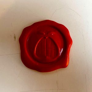 May include: A vibrant red wax seal with a central design of a stylized letter or emblem. The seal is circular with a smooth, glossy surface, and is set against a neutral background.