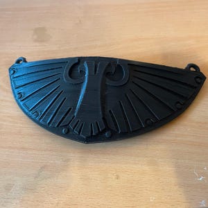 May include: A black, semi-circular decorative emblem featuring a central stylized eagle motif with symmetrical, radiating wing patterns. The piece has a textured finish and includes small circular loops at both top corners for attachment purposes.