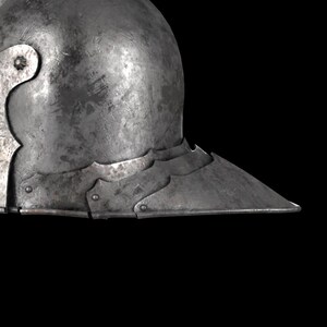 May include: A close-up of a weathered, metallic helmet with a pointed visor. The helmet has a rounded top and a series of decorative, scalloped edges along the visor. The metal shows signs of wear and age, with a dark, matte finish.