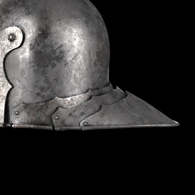 May include: A close-up of a weathered, metallic helmet with a pointed visor. The helmet has a rounded top and a series of decorative, scalloped edges along the visor. The metal shows signs of wear and age, with a dark, matte finish.