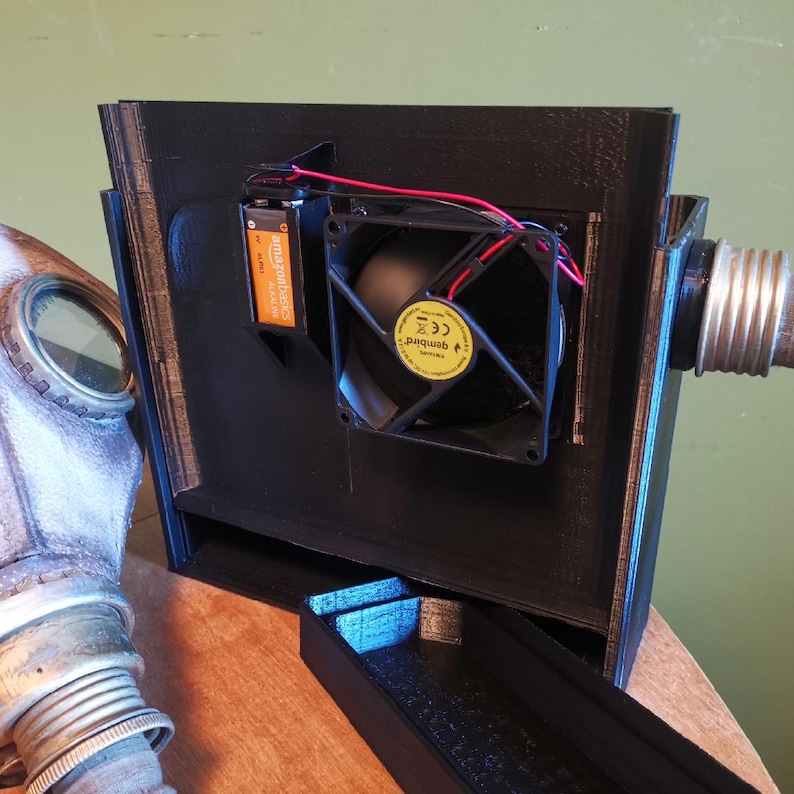 May include: A black 3D printed box with a small fan and a battery inside. The box has a small opening on the side and a hose attached to the back. The box is sitting on a wooden surface.