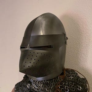 Pode incluir: Um capacete de metal cinza escuro com viseira e protetor facial perfurado. O capacete faz parte de um conjunto de armadura de cavaleiro medieval. O capacete &eacute; usado sobre uma coifa de malha.
