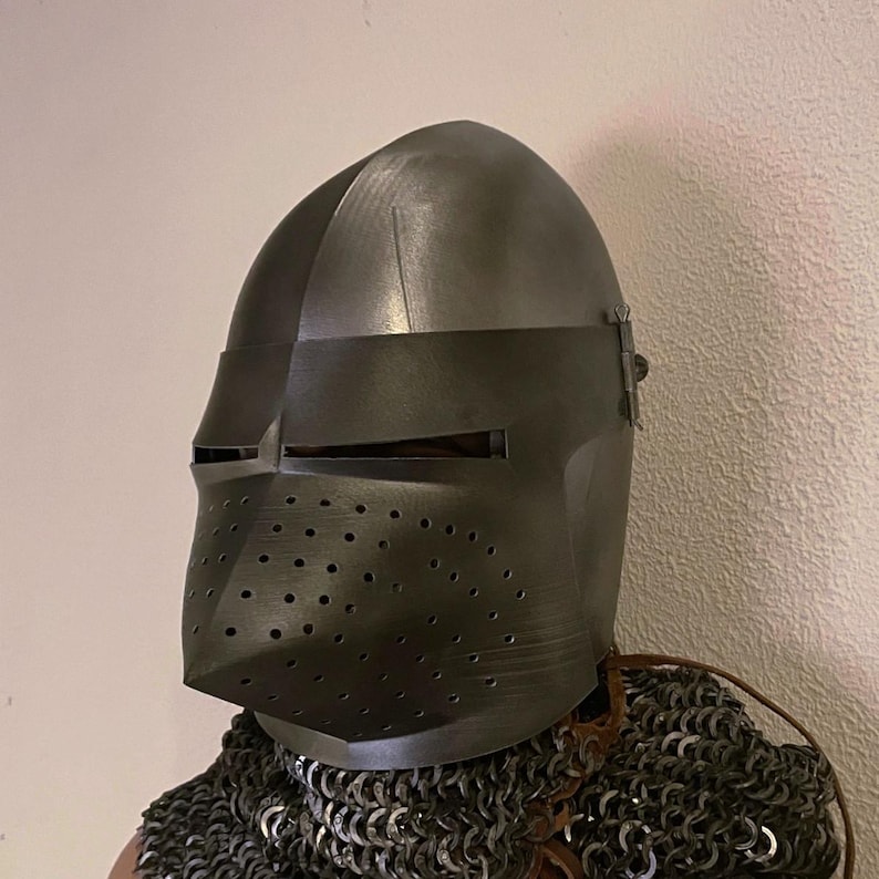 Pode incluir: Um capacete de metal cinza escuro com viseira e protetor facial perfurado. O capacete faz parte de um conjunto de armadura de cavaleiro medieval. O capacete &eacute; usado sobre uma coifa de malha.