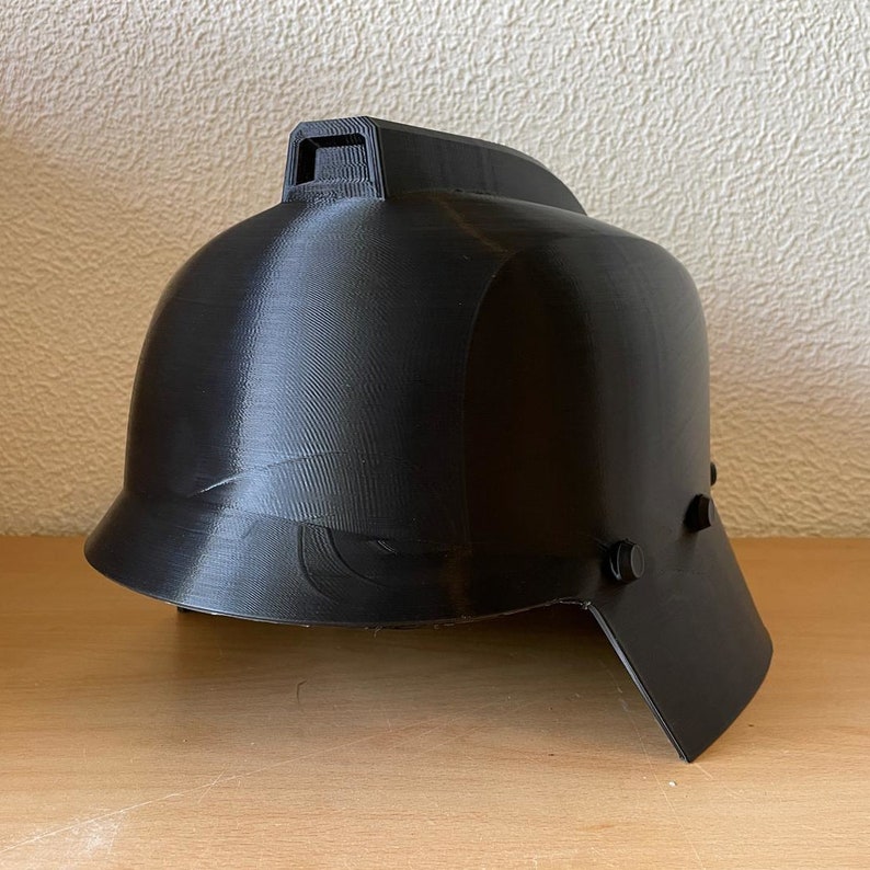May include: A black plastic helmet with a raised ridge on the top and a visor on the side. The helmet has a textured surface and is designed to protect the head.