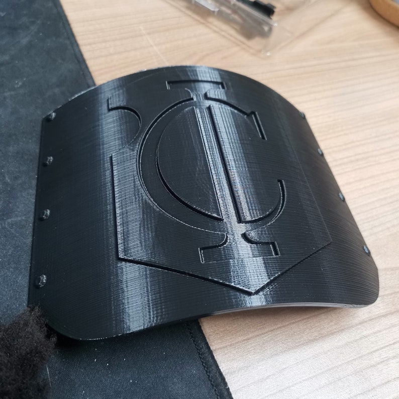 May include: A black plastic 3D printed shield with a raised, stylized letter "C" in the center.