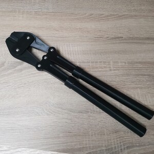 Puede incluir: Cortador de cable de metal negro con dos mangos largos. El cortador tiene una placa de metal plateado con un peque&ntilde;o agujero en el centro.