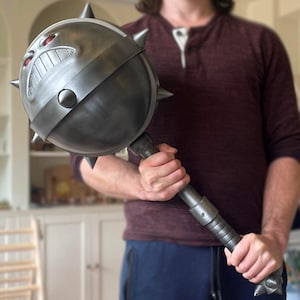 May include: A large, gray mace with a menacing face design and spiked protrusions. The mace features a round head with a detailed, angry expression and a long, textured handle. A prop for cosplay or display.