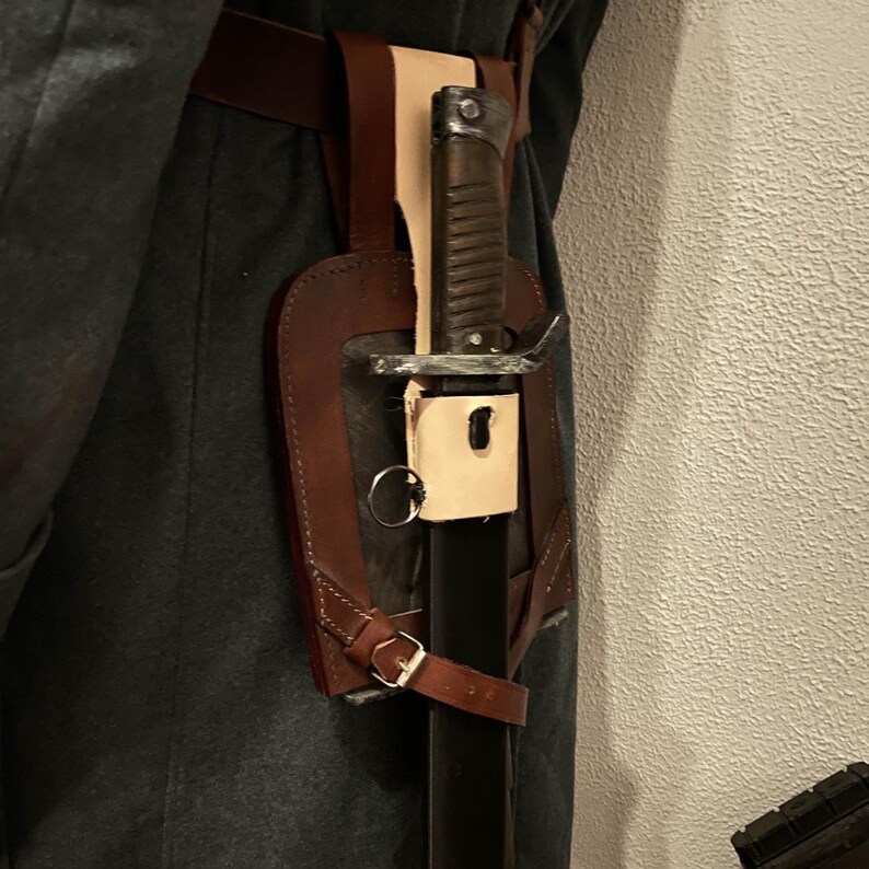 May include: A brown leather scabbard with a metal buckle and a ring. A sword is attached to the scabbard. The scabbard is worn on a belt.