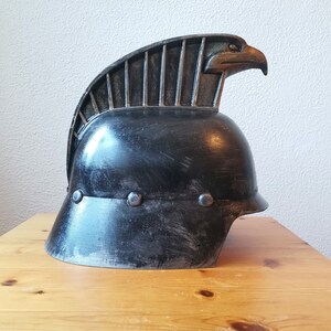 Puede incluir: Un casco negro con una cresta de &aacute;guila estilizada. El casco tiene un acabado desgastado y est&aacute; hecho de metal.