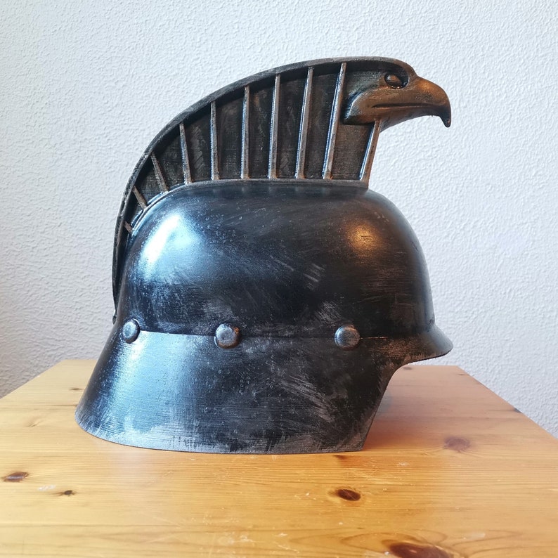 Puede incluir: Un casco negro con una cresta de &aacute;guila estilizada. El casco tiene un acabado desgastado y est&aacute; hecho de metal.