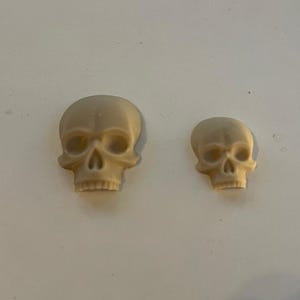 May include: Two beige ceramic skull figurines, one larger than the other.
