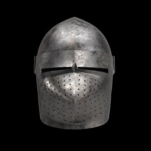 Pode incluir: Um capacete medieval de a&ccedil;o com uma parte superior arredondada e uma placa frontal com pequenos orif&iacute;cios de ventila&ccedil;&atilde;o. O capacete tem uma fenda horizontal para os olhos e uma pequena protuber&acirc;ncia pontiaguda acima. O metal tem uma apar&ecirc;ncia desgastada e envelhecida.