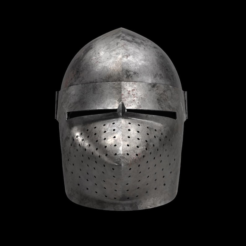 Pode incluir: Um capacete medieval de a&ccedil;o com uma parte superior arredondada e uma placa frontal com pequenos orif&iacute;cios de ventila&ccedil;&atilde;o. O capacete tem uma fenda horizontal para os olhos e uma pequena protuber&acirc;ncia pontiaguda acima. O metal tem uma apar&ecirc;ncia desgastada e envelhecida.