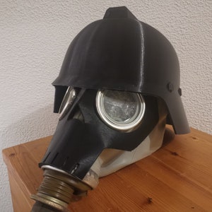 K&ouml;nnte beinhalten: Eine schwarze Gasmaske mit einem daran befestigten schwarzen Helm. Der Helm hat ein Visier und einen Kinnriemen. Die Gasmaske hat ein gro&szlig;es rundes Augenglas und einen Nasenaufsatz.