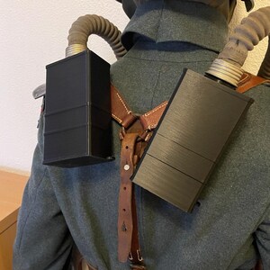 May include: Two black rectangular canteens with a brown leather strap and buckles. The canteens are worn on a person's back.