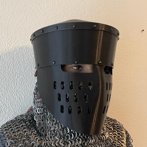 May include: A black medieval helmet with a visor featuring multiple small holes and vertical slits. The helmet is riveted and worn over chainmail armor. The person's eyes are visible through the visor.