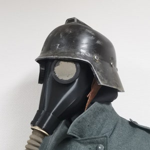 May include: A black gas mask with a clear visor attached to a black helmet with a brown leather strap. The helmet has a metal visor and a textured surface.