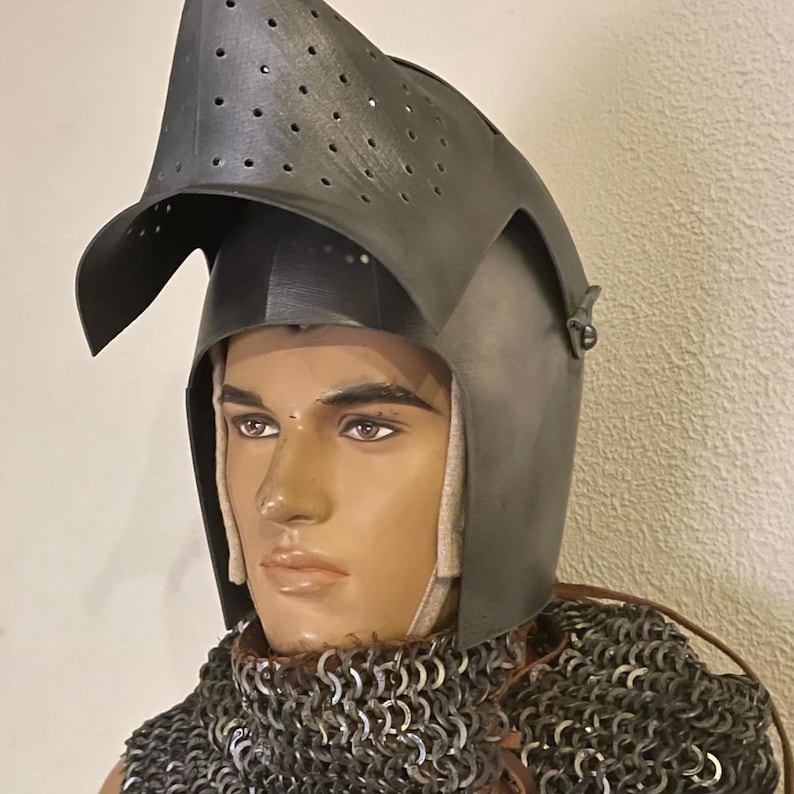 Pode incluir: Um capacete medieval cinza escuro com viseira e orif&iacute;cios de ventila&ccedil;&atilde;o, exibido em um manequim. O capacete &eacute; feito de metal e &eacute; combinado com uma gola de malha de corrente, t&iacute;pica de armaduras hist&oacute;ricas.