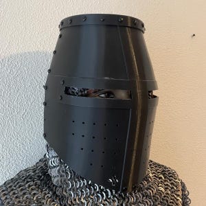 May include: A black, medieval-style helmet with a cylindrical shape and rivet detailing. The helmet has a visor with horizontal eye slits and small ventilation holes. The person wearing the helmet is also wearing chainmail.