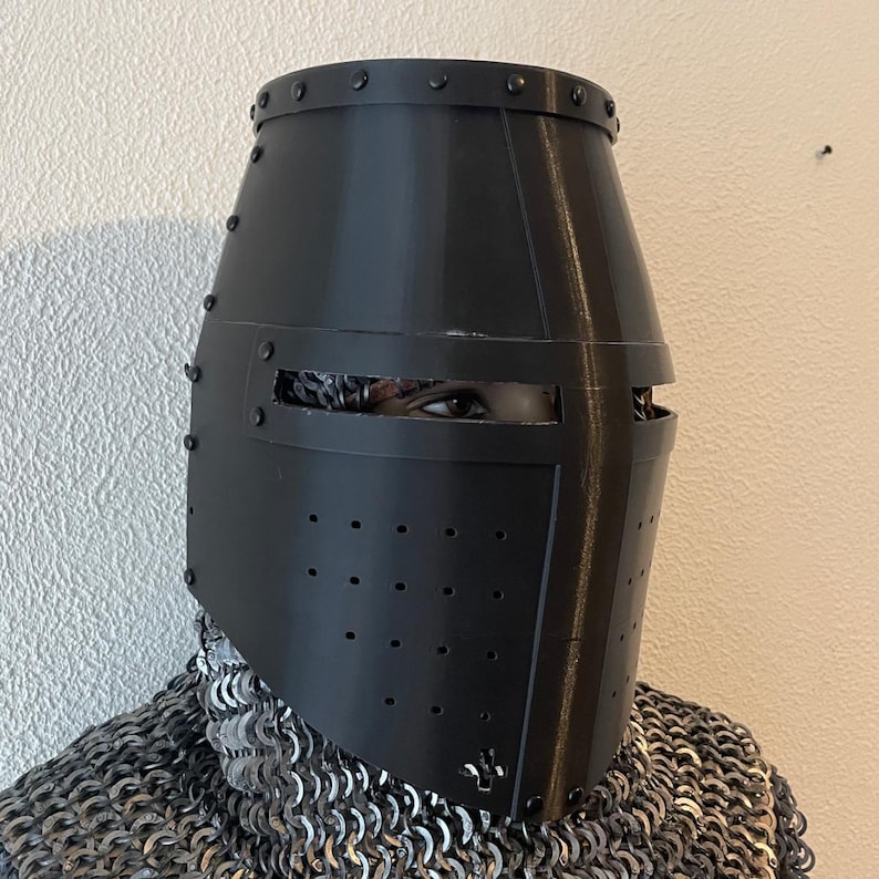 May include: A black, medieval-style helmet with a cylindrical shape and rivet detailing. The helmet has a visor with horizontal eye slits and small ventilation holes. The person wearing the helmet is also wearing chainmail.
