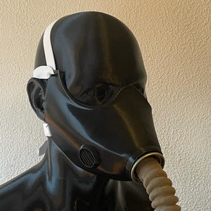 May include: A black gas mask with a white strap and a brown hose attached to the side. The mask has a round, black vent on the side.
