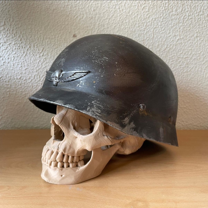 May include: A weathered gray military helmet with a skull and wings insignia sits atop a beige skull. The helmet has a worn, distressed look, adding to its vintage appeal.