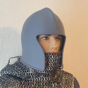 May include: A gray medieval helmet with a rounded top and face guard, paired with a chainmail coif. The helmet is displayed on a mannequin, showcasing its design and construction. The coif is made of interlocking metal rings.