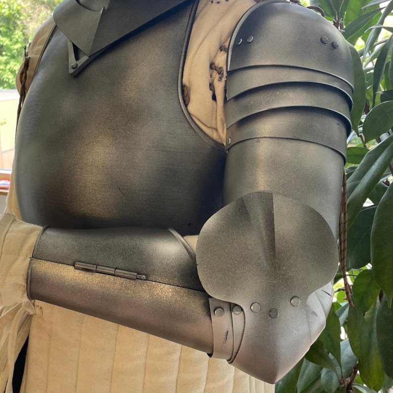 May include: A dark grey metal breastplate and articulated arm armor set worn over a cream-colored quilted garment. The armor includes a chest plate, shoulder pauldrons, and a hinged forearm guard with a prominent elbow cop featuring rivets.