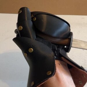 May include: A black leather gauntlet featuring articulated plates secured with brass-coloured metal rivets. The protective glove is constructed with layered segments covering the hand and wrist, and it is shown gripping a wooden-handled metal blade.
