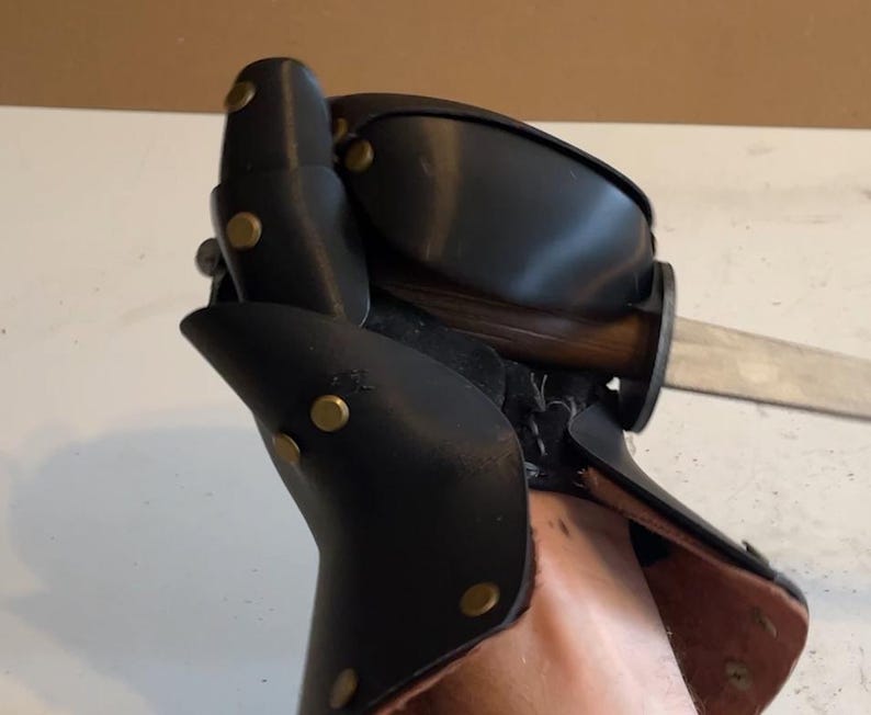 May include: A black leather gauntlet featuring articulated plates secured with brass-coloured metal rivets. The protective glove is constructed with layered segments covering the hand and wrist, and it is shown gripping a wooden-handled metal blade.
