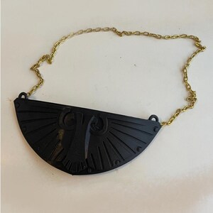 May include: A black, semi-circular pendant featuring a raised, stylized eagle emblem with radiating lines, attached to a thin, gold-toned metal chain. The pendant has a matte finish and small circular details along its curved bottom edge.