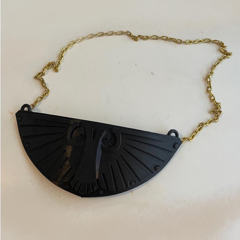 May include: A black, semi-circular pendant featuring a raised, stylized eagle emblem with radiating lines, attached to a thin, gold-toned metal chain. The pendant has a matte finish and small circular details along its curved bottom edge.