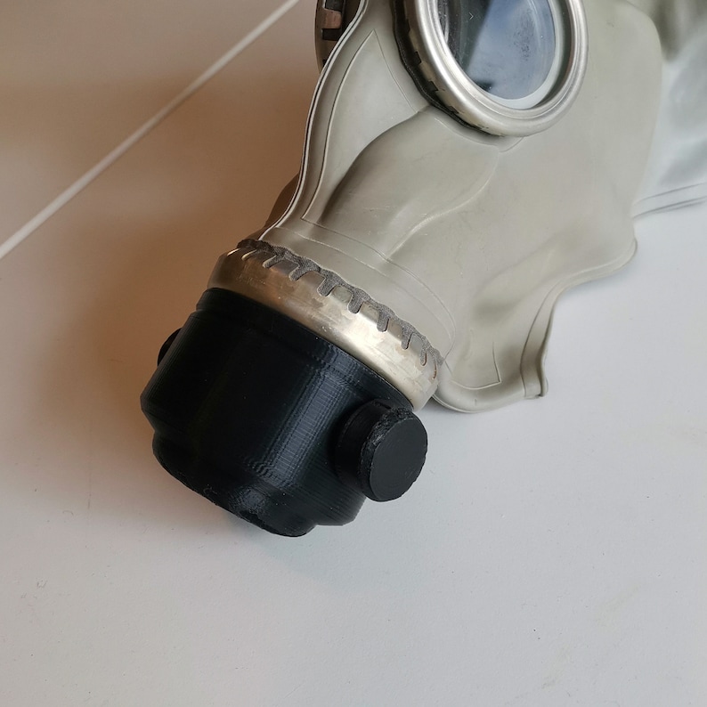 May include: A close-up of a gas mask with a black rubber mouthpiece and a silver metal ring around the edge. The mask is made of a light gray rubber.