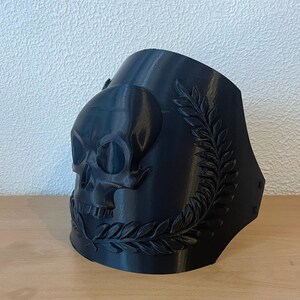 May include: A black decorative skull-shaped object with a laurel wreath design. The skull has detailed eye sockets and a nose cavity. The object is made of a solid material and has a smooth finish. The design is likely 3D printed.