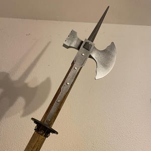 Op de afbeelding: Een middeleeuwse strijdbijl met een houten schacht, metalen blad en punt. De bijlkop heeft een gebogen blad en een hamerachtig uitsteeksel. De metalen componenten hebben een zilveren afwerking en de houten schacht is lichtbruin.