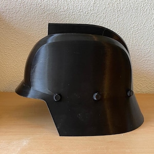 Può includere: Un casco nero con una parte superiore curva e una visiera. Il casco ha una forma arrotondata con rivetti sui lati. Il casco è realizzato in un materiale solido e opaco.