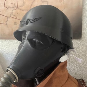May include: A black gas mask with a white strap attached to a black helmet. The helmet has a skull and wings emblem on the front.