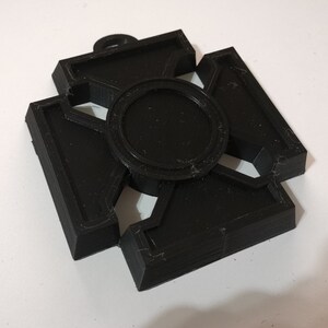 May include: A black, 3D-printed pendant with a cross-like design. The pendant features a central circular element and four rectangular arms, each with a geometric cutout. A small loop is at the top for hanging.