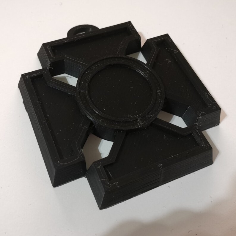 May include: A black, 3D-printed pendant with a cross-like design. The pendant features a central circular element and four rectangular arms, each with a geometric cutout. A small loop is at the top for hanging.