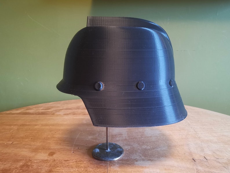 May include: A black plastic helmet with a smooth, textured surface. The helmet has a rounded top and a wide brim. There are three small, round, black accents on the side of the helmet.