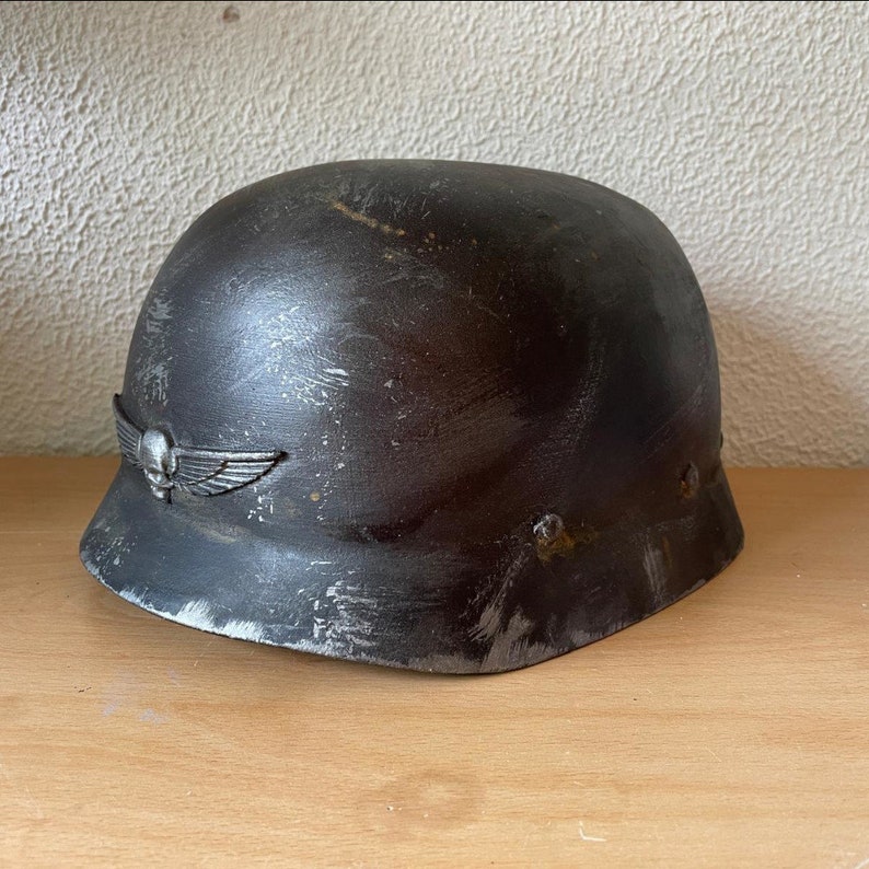 May include: A black helmet with a silver skull and wings emblem. The helmet has a distressed, weathered look and is made of metal.
