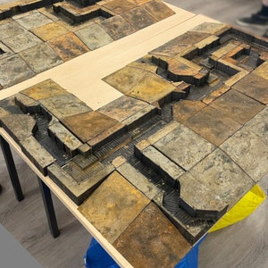 May include: A tabletop wargaming terrain set featuring a detailed, multi-level trench system made of brown and gray textured tiles. The trenches are connected by walkways and have various levels of detail, including small walls and steps.