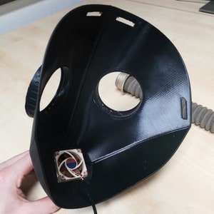 May include: A black 3D printed mask with a small fan attached to the inside. The mask has a hole for a hose and a hole for the nose.