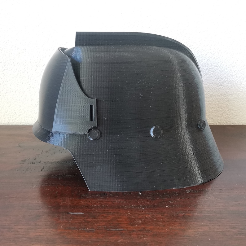 May include: A black plastic helmet with a textured surface. The helmet has a rounded top and a flat brim. There are two small holes on the side of the helmet.