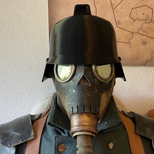 May include: A black gas mask with a skull-shaped front and two round eye pieces. The mask is attached to a brown gas mask canister with a hose. The mask is worn over a black helmet with a visor.