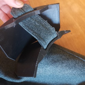 May include: A black plastic object with a gray fabric strap. The object is curved and has a textured surface.